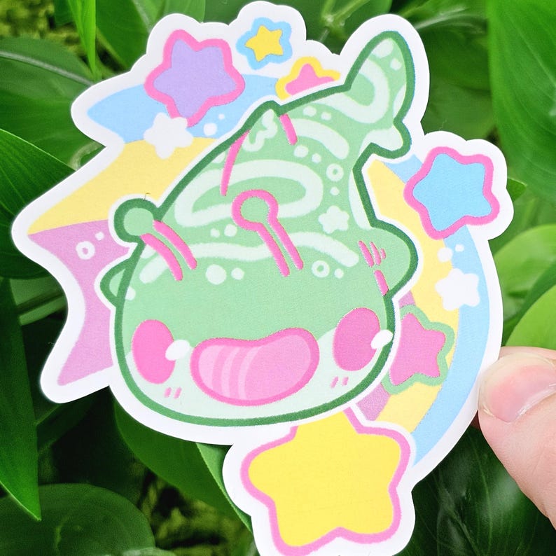 May include: A die-cut sticker featuring a cartoon alien with a green body, pink cheeks, and a smiling face. The sticker has a pastel rainbow and star design in shades of yellow, blue, pink, and purple. The sticker is held against a background of green leaves.
