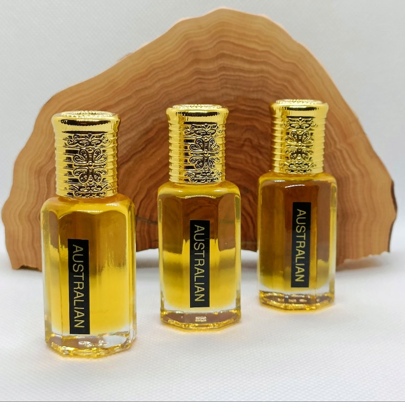 May include: Three small, clear glass bottles filled with a golden liquid, each with a gold-colored cap. The caps have an ornate design. Each bottle has a black label that reads "AUSTRALIAN". The bottles are in front of a wooden background.