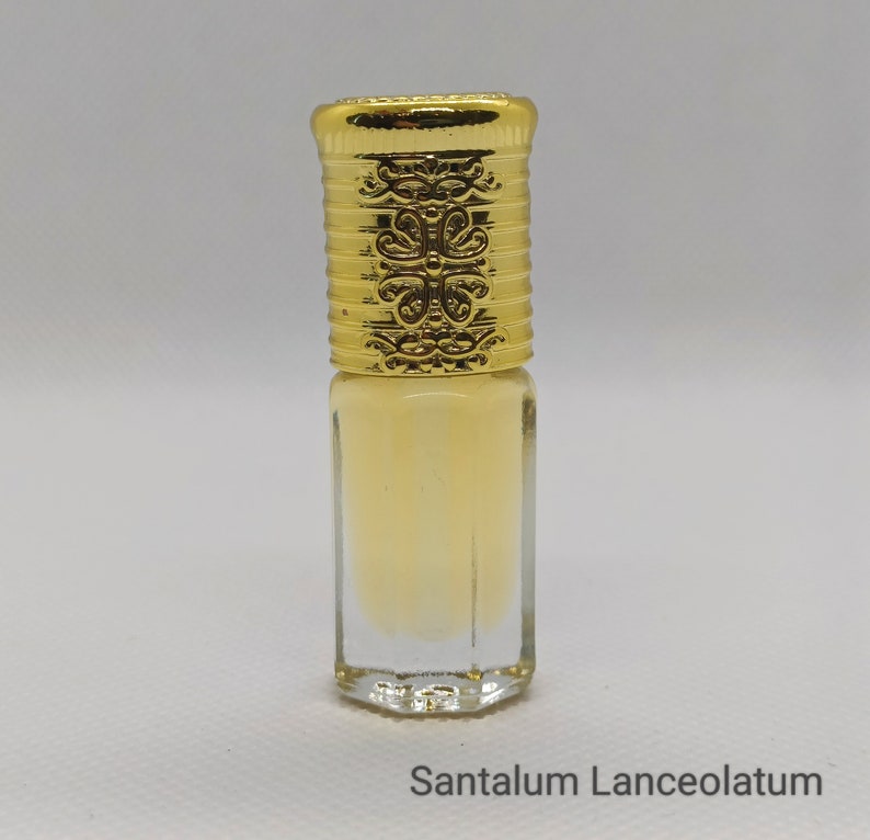 May include: A small, clear glass bottle filled with a pale yellow liquid. The bottle has a decorative gold-colored cap with ornate detailing. The text "Santalum Lanceolatum" is printed at the bottom of the image.