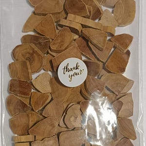 May include: A clear plastic bag filled with numerous small, irregular, light brown wooden slices. The pieces vary in shape and size, displaying natural wood grain patterns. A circular white sticker with the text thank you in gold is attached.