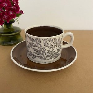 May include: A white teacup and saucer with a brown floral pattern. The teacup is filled with dark brown liquid.