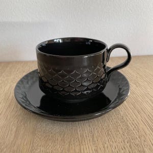 Può includere: Un set da tè nero con tazza e piattino con un motivo geometrico. La tazza e il piattino sono in ceramica e hanno una finitura lucida.