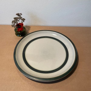 May include: A large, white ceramic dinner plate with a green rim and two concentric green bands. The plate is on a brown surface.