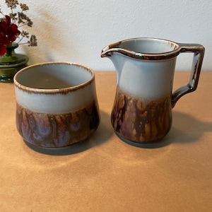 May include: A brown and white ceramic creamer and sugar bowl set with a mottled glaze. The creamer has a handle and a spout. The sugar bowl is round and has a wide opening.