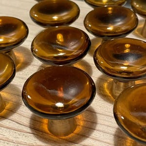 Puede incluir: Una colección de pomos redondos para armarios con tapas de vidrio translúcido de color ámbar y superficie lisa. Cada pomo está montado sobre una base metálica cilíndrica con acabado pulido, creando una apariencia uniforme en todo el conjunto.