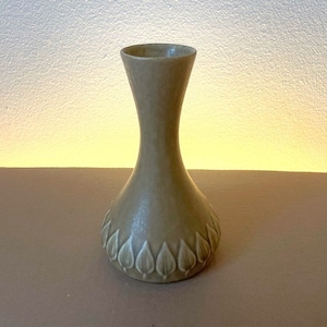 Può includere: Un piccolo vaso in ceramica marrone chiaro con collo stretto e base larga e arrotondata. Il vaso è decorato con un motivo ripetitivo di foglie bianche intorno alla base.