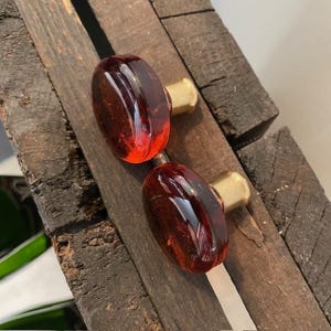 Könnte beinhalten: Zwei ovale Schrankknöpfe mit einem tiefroten Glaskörper und einem goldfarbenen Metallsockel. Die Knöpfe werden auf einer rustikalen Holzoberfläche präsentiert, die ihr Design und ihre Farbe hervorhebt.