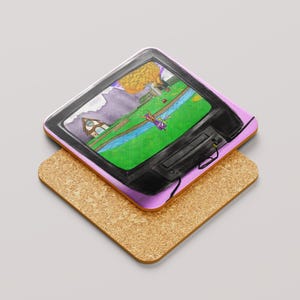May include: A square coaster with a hand-drawn image of a retro television showing a video game scene. The coaster has a pink border and sits on a cork coaster. The TV screen displays a green field, a river, and a house.