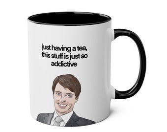 May include: White ceramic mug with a black interior and handle. The mug features an illustration of a smiling person in a suit and tie, with the text "just having a tea, this stuff is just so addictive."