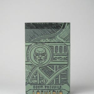 May include: A rectangular sage green box with a textured, embossed surface featuring a stylized face within a circular frame, geometric lines, and the text "DOOM PREVAILS." and "LATVERIA" printed in a dark, bold font at the bottom of the front panel.