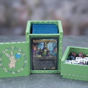 May include: A light green wooden deck box with a removable lid featuring a painted rabbit holding a blue flower. The box displays a Ms. Bumbleflower trading card in a clear front slot and contains a separate compartment filled with polyhedral dice.