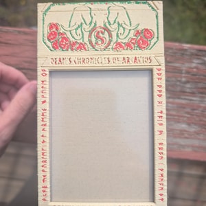 May include: Rectangular vintage card with a cream-colored background. The top features two green elephants, red roses, and the text "DEAN'S CHRONICLES OF ARCAVIOS". The frame is bordered with red text. The card is held by a person.