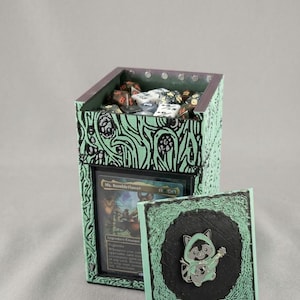 May include: A mint green and black card and dice holder with a decorative floral design. The box contains dice and a card that reads "Ms. Bumbleflower". A matching pin with a raccoon playing a ukulele is also visible.