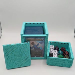 May include: A teal-colored card game storage set. The set includes a box for cards, a small box for dice, and a lid. The dice box has the text "MATH IS THE MAGIC AT THE HEART OF NATURE." A card is visible inside the card box.