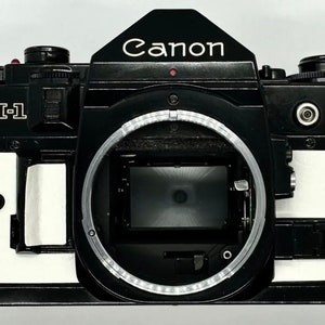 May include: A black Canon A-1 film camera with a white leatherette covering. The camera has a silver lens ring and a red dot on the top.
