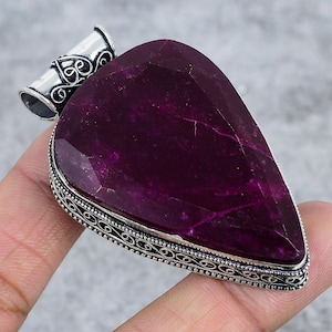 May include: A large, teardrop-shaped pendant featuring a deep magenta gemstone. The stone is faceted and set in a silver-toned metal frame with an ornate, filigree design. The bail is also silver-toned with decorative swirls.