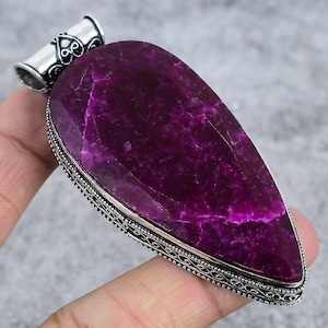 May include: A large, teardrop-shaped pendant featuring a deep magenta gemstone. The stone is set in a silver-toned metal frame with detailed filigree. The pendant has a decorative bail for a necklace.