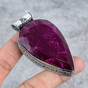Pode incluir: Um pingente grande em forma de gota com uma pedra preciosa magenta escura. A pedra é facetada e colocada em uma moldura de metal prateado com detalhes intrincados. A argola é um tubo prateado com redemoinhos decorativos.