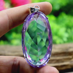Op de afbeelding: Een ovale hanger met een grote, facetgeslepen edelsteen. De edelsteen toont een kleurengradi&euml;nt van paars, groen en roze. De hanger is gezet in een zilverkleurig metalen frame en heeft een kleine lus voor een ketting.