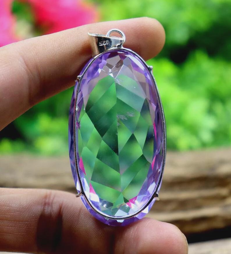 Op de afbeelding: Een ovale hanger met een grote, facetgeslepen edelsteen. De edelsteen toont een kleurengradi&euml;nt van paars, groen en roze. De hanger is gezet in een zilverkleurig metalen frame en heeft een kleine lus voor een ketting.