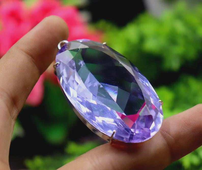 Op de afbeelding: Een grote, facetgeslepen, ovaalvormige edelsteen met een zilverkleurige metalen zetting. De edelsteen is voornamelijk lavendelkleurig met groene en roze flitsen, vastgehouden tussen vingers tegen een vage roze en groene achtergrond.