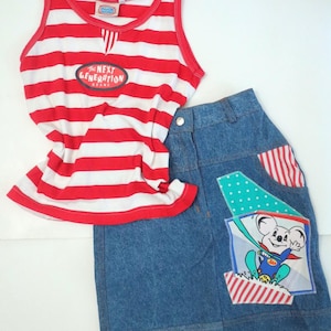May include: A red and white striped tank top with the text "The Next Generation Brand" on the front. A blue denim skirt with a red, white and blue patchwork design featuring a cartoon koala wearing a superhero costume.