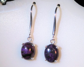 Puede incluir: Pendientes colgantes con piedras preciosas ovaladas de color púrpura intenso con vetas negras, engastadas en metal plateado. Los pendientes tienen largos ganchos plateados curvados para orejas perforadas.