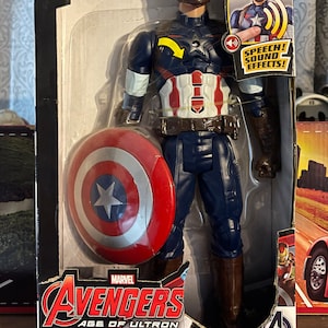 Puede incluir: Figura de acci&oacute;n de Capit&aacute;n Am&eacute;rica Titan Hero Tech en su embalaje. La figura lleva un traje azul, blanco y rojo y sostiene un escudo. El embalaje incluye el texto "Avengers Age of Ultron" y "Speech! Sound Effects!"