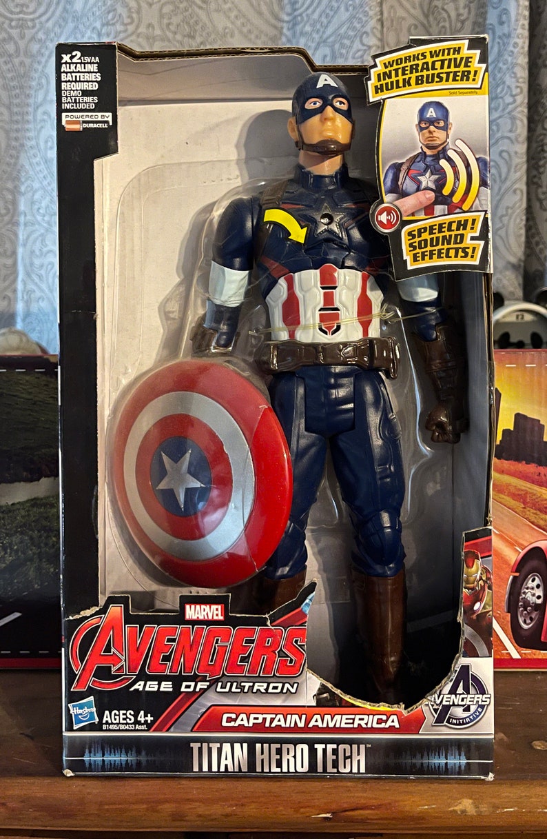 Puede incluir: Figura de acci&oacute;n de Capit&aacute;n Am&eacute;rica Titan Hero Tech en su embalaje. La figura lleva un traje azul, blanco y rojo y sostiene un escudo. El embalaje incluye el texto "Avengers Age of Ultron" y "Speech! Sound Effects!"