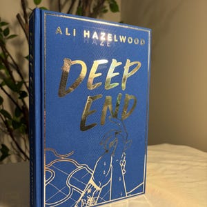 May include: A hardcover book titled Deep End by Ali Hazelwood with a vibrant blue cover. The title and a graphic illustration of two hands reaching toward each other are printed in a reflective gold foil. The spine also features the title and author.