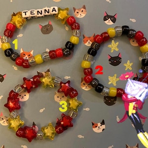 May include: Three beaded bracelets with red, yellow, black, and clear beads. One bracelet spells "TENNA" with white letter beads. The bracelets feature star-shaped and round beads. The background is light blue with cat illustrations.