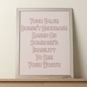 Puede incluir: Una impresión enmarcada con un fondo rosa y texto blanco que dice "Tu valor no disminuye en función de la incapacidad de alguien para ver tu valor". El marco es de madera clara.