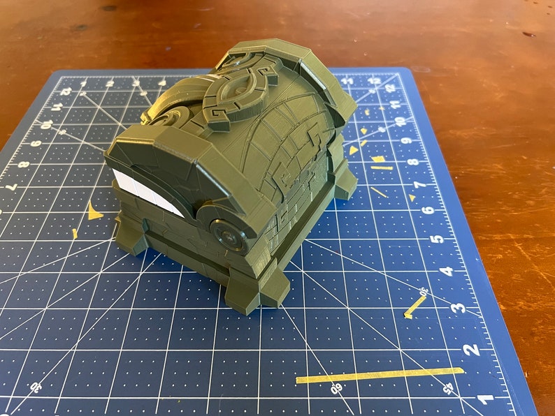May include: A green 3D printed treasure chest with intricate details and a white sticker on the front.