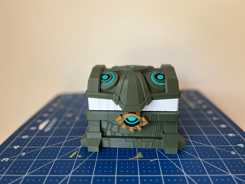 May include: A green and white treasure chest with a gold and blue eye design. The chest is made of plastic and has a detailed design.