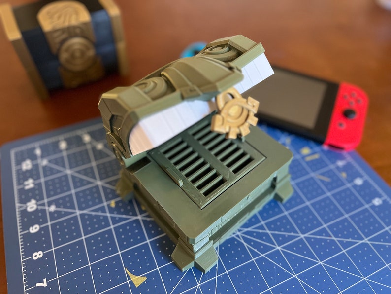 May include: A green and gold 3D printed box with a hinged lid, a gear-shaped handle, and a decorative metal grate. The box is open and the lid is propped up.