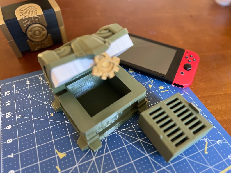 May include: A green, 3D printed treasure chest with a hinged lid, open to reveal an empty interior. The chest has intricate details and a gear-like design on the lid. A smaller, rectangular, green, 3D printed piece with a grid pattern is also visible.