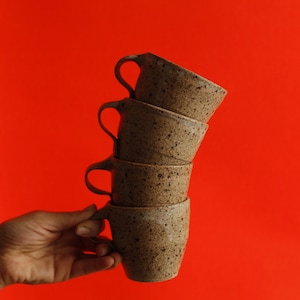 May include: A hand holding a stack of four brown speckled ceramic mugs. The mugs are stacked in a pyramid shape, with the largest mug at the bottom and the smallest mug at the top. The mugs are all slightly different shades of brown, and they have a speckled glaze.