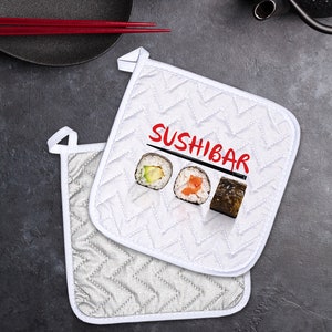 May include: Two white quilted pot holders with a red "SUSHIBAR" text and a graphic of three sushi rolls. The pot holders have a white border and a chevron pattern.