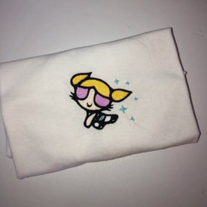 May include: White t-shirt with an embroidered image of a cartoon character with blonde hair, pink sunglasses, and a blue outfit. The character is surrounded by blue stars.