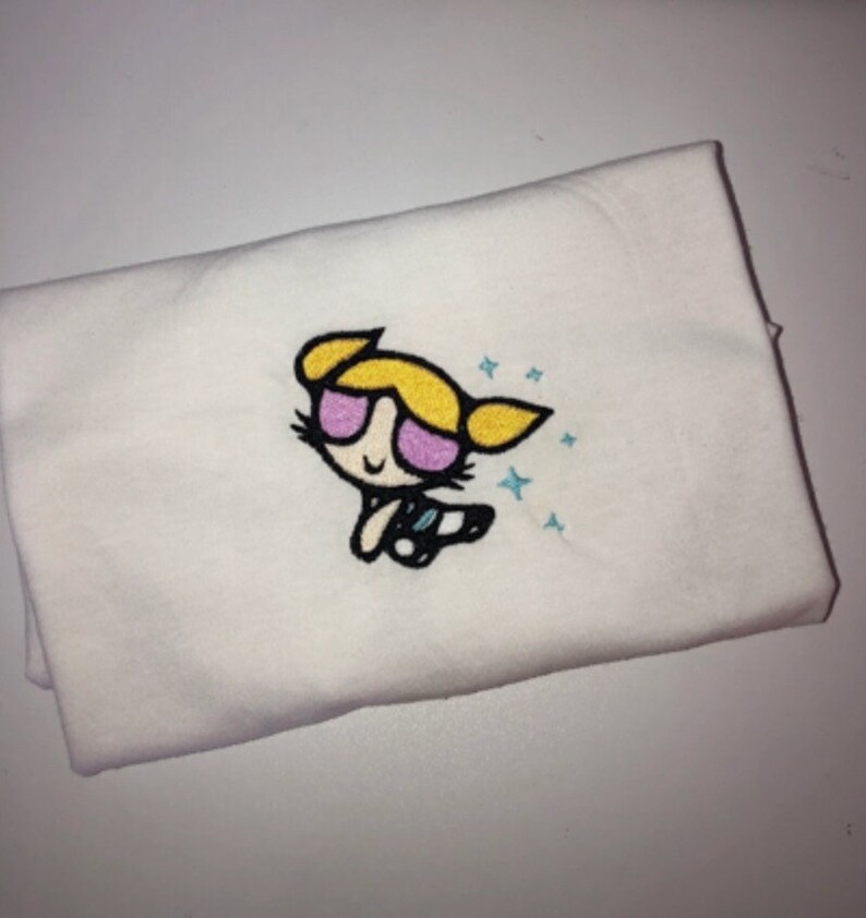 May include: White t-shirt with an embroidered image of a cartoon character with blonde hair, pink sunglasses, and a blue outfit. The character is surrounded by blue stars.
