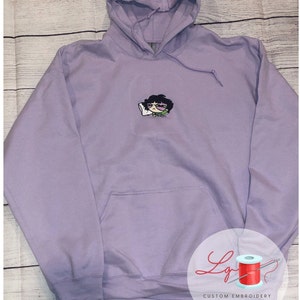May include: A lavender hoodie with a small embroidered cartoon character wearing sunglasses and holding a pillow. The hoodie has a pouch pocket and a drawstring hood.