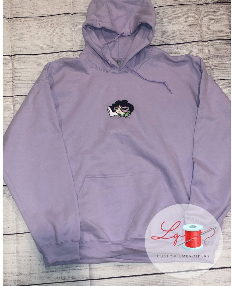May include: A lavender hoodie with a small embroidered cartoon character wearing sunglasses and holding a pillow. The hoodie has a pouch pocket and a drawstring hood.
