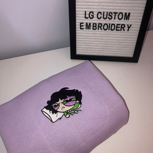 May include: Purple sweatshirt with an embroidered image of a cartoon character with green eyes and black hair, sleeping on a white pillow. The text "LG CUSTOM EMBROIDERY" is on a white letter board in a black frame.