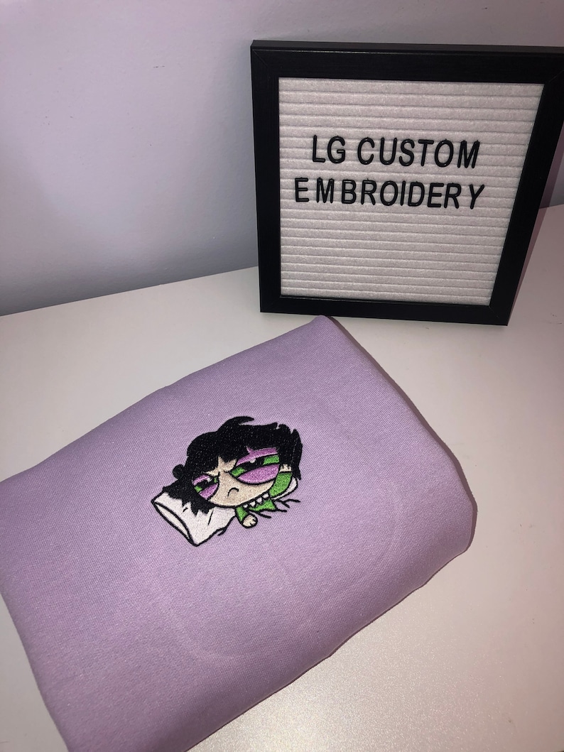 May include: Purple sweatshirt with an embroidered image of a cartoon character with green eyes and black hair, sleeping on a white pillow. The text "LG CUSTOM EMBROIDERY" is on a white letter board in a black frame.