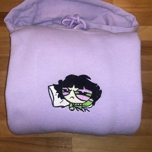 May include: A lavender hoodie with an embroidered cartoon character of a girl with black hair, green eyes, and a pink face, sleeping on a white pillow. The character is wearing a green shirt.