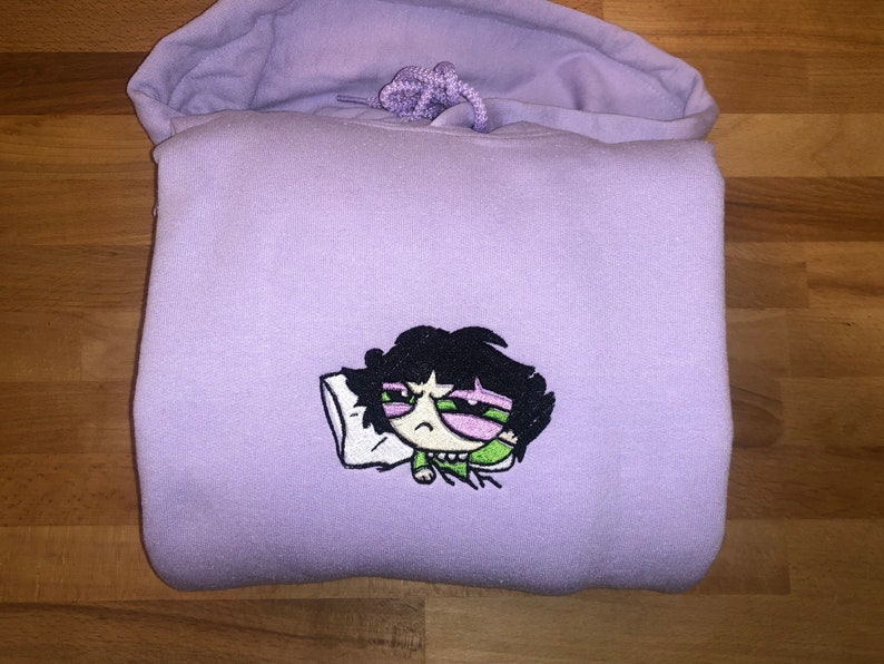 May include: A lavender hoodie with an embroidered cartoon character of a girl with black hair, green eyes, and a pink face, sleeping on a white pillow. The character is wearing a green shirt.