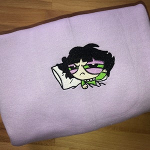 May include: A purple sweatshirt with an embroidered image of a cartoon character with black hair, green eyes, and a pink face. The character is lying down with a pillow and appears to be sleeping.
