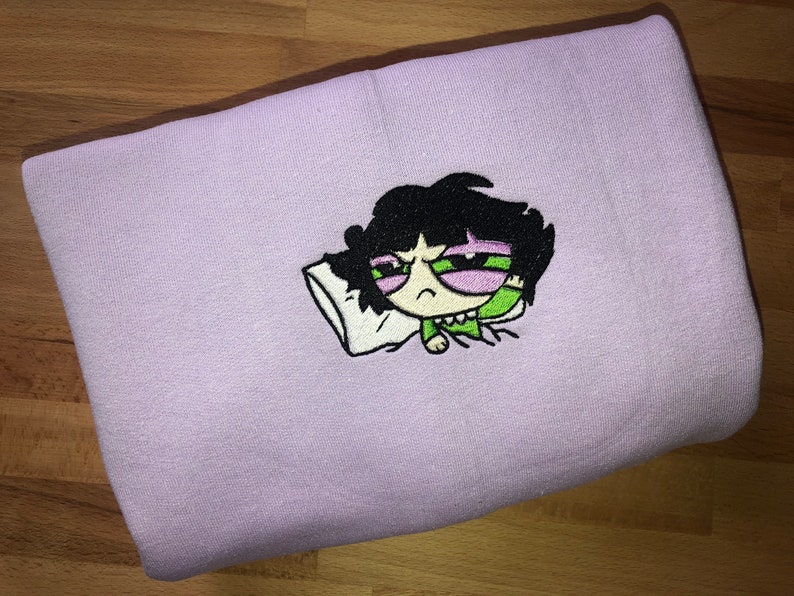May include: A purple sweatshirt with an embroidered image of a cartoon character with black hair, green eyes, and a pink face. The character is lying down with a pillow and appears to be sleeping.