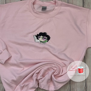 May include: Pink sweatshirt with an embroidered cartoon character of a girl with black hair, green eyes, and a green shirt, sleeping on a white pillow. The character has a black eye.