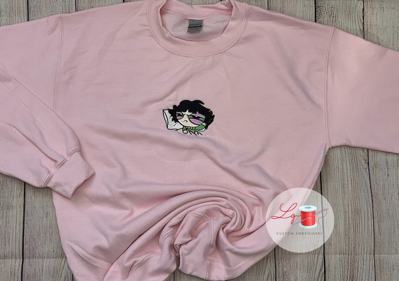 May include: Pink sweatshirt with an embroidered cartoon character of a girl with black hair, green eyes, and a green shirt, sleeping on a white pillow. The character has a black eye.